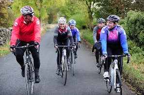 A group of five cyclists are riding on a country road surrounded by lush greenery. They are all wearing helmets and cycling attire, indicating they are on a casual group ride. Their expressions are cheerful, suggesting enjoyment and camaraderie during the activity.