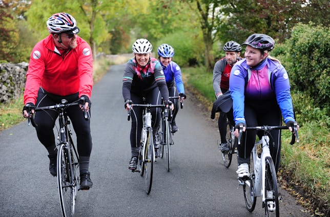 A group of five cyclists are riding on a country road surrounded by lush greenery. They are all wearing helmets and cycling attire, indicating they are on a casual group ride. Their expressions are cheerful, suggesting enjoyment and camaraderie during the activity.