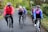 A group of five cyclists are riding on a country road surrounded by lush greenery. They are all wearing helmets and cycling attire, indicating they are on a casual group ride. Their expressions are cheerful, suggesting enjoyment and camaraderie during the activity.