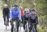 A group of cyclists is riding along a paved road, surrounded by lush greenery. The cyclists are wearing colorful helmets and clothing, and they appear to be enjoying the ride. Sunlight filters through the surrounding trees, creating a cheerful atmosphere.