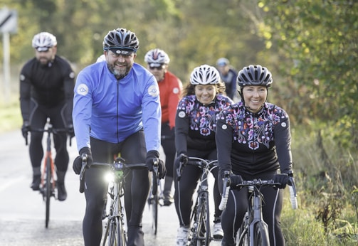 A group of cyclists is riding along a paved road, surrounded by lush greenery. The cyclists are wearing colorful helmets and clothing, and they appear to be enjoying the ride. Sunlight filters through the surrounding trees, creating a cheerful atmosphere.
