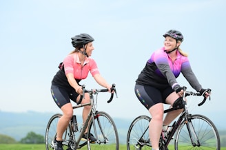 Two women are riding bicycles outdoors in a scenic area. They are wearing cycling gear including helmets and sports jerseys, one in pink and black and the other in purple and black. The background features a clear sky and rolling hills, indicating they are cycling in a rural or natural setting.