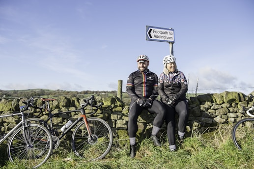 Two cyclists wearing helmets and cycling attire sit on a stone wall beside a footpath sign that reads 'Footpath to Addingham.' Their bicycles are propped against the wall on either side. The background features an open field with hills under a clear blue sky.