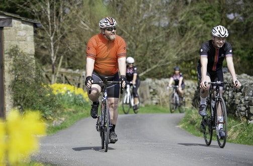 A group of cyclists is riding on a rural road lined with stone walls and colorful flowers. One cyclist in an orange jersey is beside another wearing a black jersey with colored stripes. The background includes trees and a small stone structure.