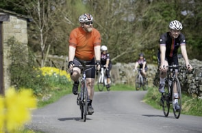 A group of cyclists is riding on a rural road lined with stone walls and colorful flowers. One cyclist in an orange jersey is beside another wearing a black jersey with colored stripes. The background includes trees and a small stone structure.