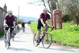A group of cyclists wearing matching outfits rides along a sunny rural road. They are cycling past an old-fashioned red telephone booth situated on a grassy verge with yellow flowers. The background includes a stone building and leafless trees, suggesting springtime.