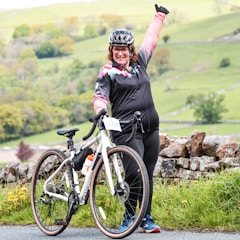 A person wearing cycling gear stands next to a bicycle with a number tag, raising one arm in a triumphant pose. The background features a scenic landscape with rolling green hills and stone walls.