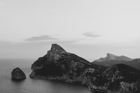 A grayscale image capturing a rugged coastline with prominent rocky cliffs jutting into the sea. The ocean surrounds the cliffs, creating a stark contrast against the dark rocky surfaces. The sky is overcast, with soft clouds adding a serene atmosphere to the scene.