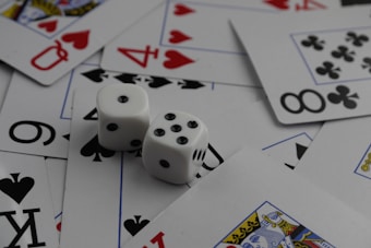A collection of playing cards spread out, featuring various suits such as hearts, clubs, and spades. Two white dice with black dots are placed prominently on top of the cards.