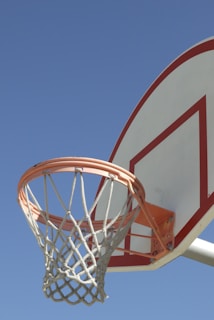 A basketball hoop and backboard with a clear blue sky background. The hoop is circular with a white net, and the backboard has a red outline.