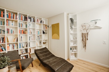 A cozy reading nook with a modern aesthetic, featuring a large white bookshelf filled with a variety of books. A comfortable gray chaise lounge is positioned alongside the bookshelf. The room includes a small plant on a side table, a dreamcatcher hanging on the wall, and a decorative shelf with small items and a potted plant. A piece of abstract wall art adds a touch of color.