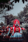 A group of people wearing traditional, colorful costumes and large ornate masks, performing in an outdoor setting surrounded by trees. The costumes feature intricate patterns with dominant colors of red, black, and white. The scene captures a cultural or ceremonial event.
