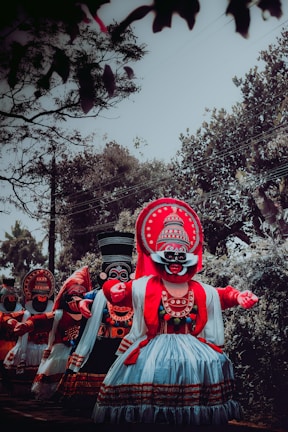 A group of people wearing traditional, colorful costumes and large ornate masks, performing in an outdoor setting surrounded by trees. The costumes feature intricate patterns with dominant colors of red, black, and white. The scene captures a cultural or ceremonial event.