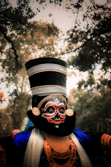 A person is wearing traditional makeup and costume typical of an Indian classical dance form. The face is painted with intricate designs in black, red, and white. They are adorned with a tall, ornate headdress and large, round earrings. The costume is richly decorated with blue fabric and gold accessories. The background is a blurred view of trees and nature.
