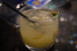 A glass filled with a light-colored cocktail containing ice cubes and garnished with a small piece of fruit. A black straw is placed inside the drink. The background is softly blurred, with bokeh lights creating a relaxed ambiance.