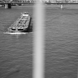 A large cargo ship travels along a wide river, creating small waves in its wake. The bridge structure in the background spans across the river, connecting urban buildings on either side. A vertical blur or obstruction bisects the image, partially obscuring the central view.
