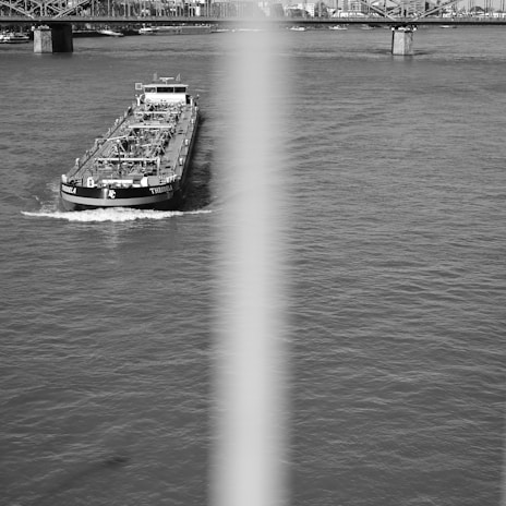 A large cargo ship travels along a wide river, creating small waves in its wake. The bridge structure in the background spans across the river, connecting urban buildings on either side. A vertical blur or obstruction bisects the image, partially obscuring the central view.