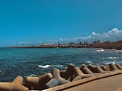 A coastal scene with a concrete breakwater in the foreground, blue ocean waves, and an industrial area with cranes along the shoreline. The sky is clear with a few scattered clouds, and the horizon shows a line of industrial structures.