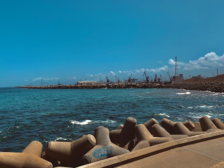 A coastal scene with a concrete breakwater in the foreground, blue ocean waves, and an industrial area with cranes along the shoreline. The sky is clear with a few scattered clouds, and the horizon shows a line of industrial structures.