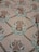 A patterned fabric with an ornate floral and geometric design. The design features symmetrical arrangements of flowers in a medallion style, surrounded by swirling decorative elements. The background consists of a subtle grid-like pattern.
