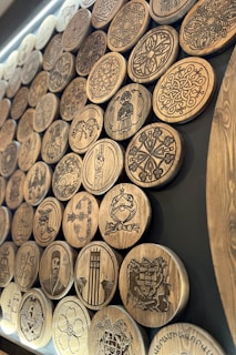 A collection of intricately carved wooden discs displayed on a dark background. The discs feature various patterns, symbols, and figures, including geometric designs, floral motifs, human figures, and animals. The carvings are detailed and showcase a range of artistic styles.