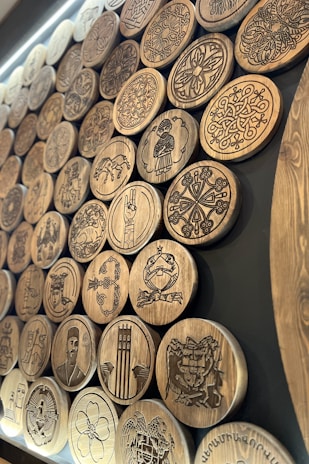 A collection of intricately carved wooden discs displayed on a dark background. The discs feature various patterns, symbols, and figures, including geometric designs, floral motifs, human figures, and animals. The carvings are detailed and showcase a range of artistic styles.