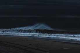 A dark and atmospheric seascape with strong waves crashing onto a deserted beach. The sky is overcast, adding to the moody and intense feel of the scene.
