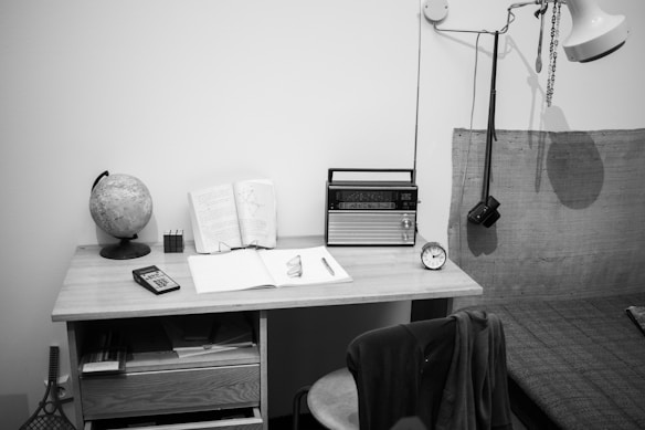 A monochromatic photograph captures a minimalist workspace. A globe, a small notebook, an open book, and a pair of reading glasses are placed on a wooden desk. A vintage radio and a small clock are also positioned on the desk. An overhead lamp hangs above, casting a shadow on the wall. Below the desk, shelves are filled with books and papers. Next to the desk, there is a chair with a coat draped over it.
