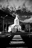 A white church with a tall steeple stands in front of a mountainous landscape under a sky filled with scattered clouds. A pathway leads up to the church from a road with a crosswalk. Street lamps border the road, and trees surround the church.