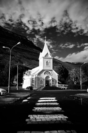 A white church with a tall steeple stands in front of a mountainous landscape under a sky filled with scattered clouds. A pathway leads up to the church from a road with a crosswalk. Street lamps border the road, and trees surround the church.