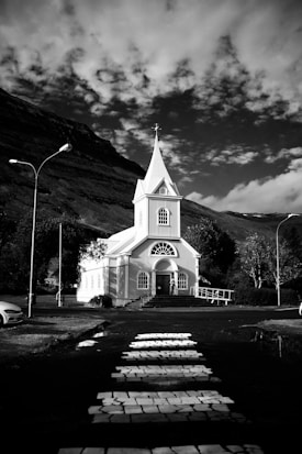 A white church with a tall steeple stands in front of a mountainous landscape under a sky filled with scattered clouds. A pathway leads up to the church from a road with a crosswalk. Street lamps border the road, and trees surround the church.