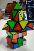 A stack of colorful puzzle cubes and pyramids with various shapes and colors arranged on a table. The background includes parts of a laptop and the blurred figure of a person using a computer.