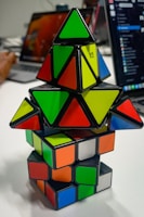 A stack of colorful puzzle cubes and pyramids with various shapes and colors arranged on a table. The background includes parts of a laptop and the blurred figure of a person using a computer.