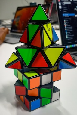 A stack of colorful puzzle cubes and pyramids with various shapes and colors arranged on a table. The background includes parts of a laptop and the blurred figure of a person using a computer.