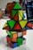 A stack of colorful puzzle cubes and pyramids with various shapes and colors arranged on a table. The background includes parts of a laptop and the blurred figure of a person using a computer.