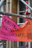 A brightly colored banner in German is attached to scaffolding. The banner has a pink and orange background with bold black text. It promotes a campaign against violence with a reference to '16 Tage gegen Gewalt an Frauen' and includes dates.
