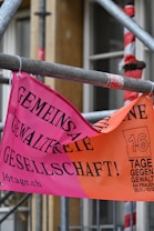 A brightly colored banner in German is attached to scaffolding. The banner has a pink and orange background with bold black text. It promotes a campaign against violence with a reference to '16 Tage gegen Gewalt an Frauen' and includes dates.