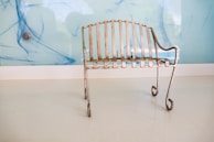 A small, rustic metal bench with a patina finish sits on a smooth, white floor. Its design features vertical slats and curling legs, and it is positioned against a light blue wall with abstract blue streaks and a metallic circular object.