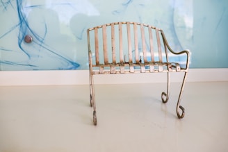 A small, rustic metal bench with a patina finish sits on a smooth, white floor. Its design features vertical slats and curling legs, and it is positioned against a light blue wall with abstract blue streaks and a metallic circular object.