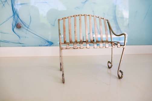 A small, rustic metal bench with a patina finish sits on a smooth, white floor. Its design features vertical slats and curling legs, and it is positioned against a light blue wall with abstract blue streaks and a metallic circular object.