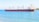 A large cargo ship is floating on calm blue waters near a shoreline. The ship has a black hull with a red bottom and features multiple cranes on its deck. There is a small green buoy in the foreground and a faint outline of land with sparse vegetation can be seen in the distance under clear skies.