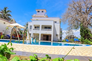 A modern, multi-story white building with a large swimming pool in the foreground sits under a bright blue sky. The pool is surrounded by a tiled deck with greenery and a few deck chairs. Umbrellas provide shade for seating areas. The atmosphere is calm and inviting, suggesting a leisurely outdoor setting.