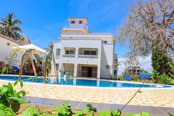 A modern, multi-story white building with a large swimming pool in the foreground sits under a bright blue sky. The pool is surrounded by a tiled deck with greenery and a few deck chairs. Umbrellas provide shade for seating areas. The atmosphere is calm and inviting, suggesting a leisurely outdoor setting.