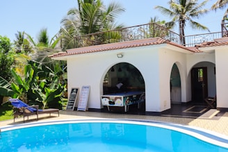 A serene outdoor setting featuring a small, white building with arched openings and a tiled roof. The area includes a blue swimming pool bordered by a tiled deck. Lush greenery and palm trees surround the scene, and a wooden lounge chair with blue cushions is visible alongside the pool. Signboards and a pool table are placed near the open area of the building.