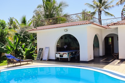 A serene outdoor setting featuring a small, white building with arched openings and a tiled roof. The area includes a blue swimming pool bordered by a tiled deck. Lush greenery and palm trees surround the scene, and a wooden lounge chair with blue cushions is visible alongside the pool. Signboards and a pool table are placed near the open area of the building.