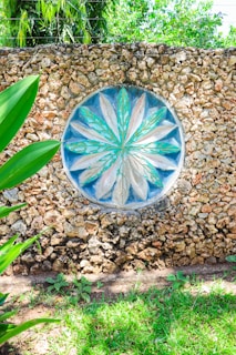 A decorative stone wall features a circular mosaic with a floral pattern in shades of turquoise and beige at its center. The texture of the rough stones contrasts with the smooth, painted design. Surrounding the wall, lush green foliage and grass add a natural frame, with sunlight casting bright highlights.