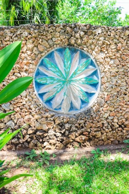 A decorative stone wall features a circular mosaic with a floral pattern in shades of turquoise and beige at its center. The texture of the rough stones contrasts with the smooth, painted design. Surrounding the wall, lush green foliage and grass add a natural frame, with sunlight casting bright highlights.