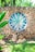 A decorative stone wall features a circular mosaic with a floral pattern in shades of turquoise and beige at its center. The texture of the rough stones contrasts with the smooth, painted design. Surrounding the wall, lush green foliage and grass add a natural frame, with sunlight casting bright highlights.