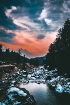 A serene landscape featuring a river flowing through a rocky terrain surrounded by dense evergreen trees. The sky is dramatic with a gradient of warm orange hues blending into cooler blues and whites, creating a picturesque backdrop.