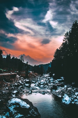 A serene landscape featuring a river flowing through a rocky terrain surrounded by dense evergreen trees. The sky is dramatic with a gradient of warm orange hues blending into cooler blues and whites, creating a picturesque backdrop.